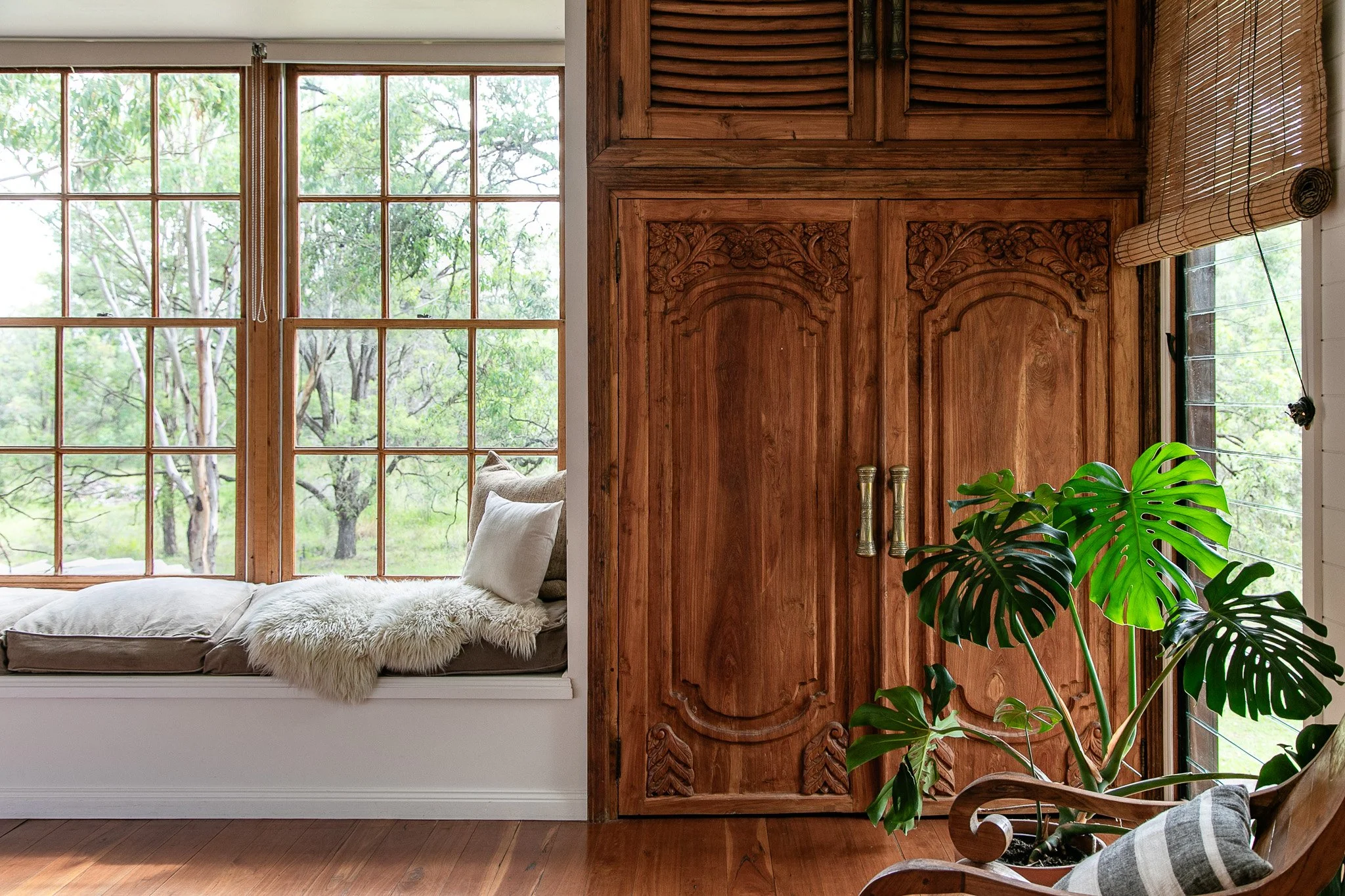 Cozy interior corner with window seat, plush pillows, soft throw, and a large leafy plant beside a carved wooden door and a window with bamboo blinds, overlooking a green outdoor landscape.