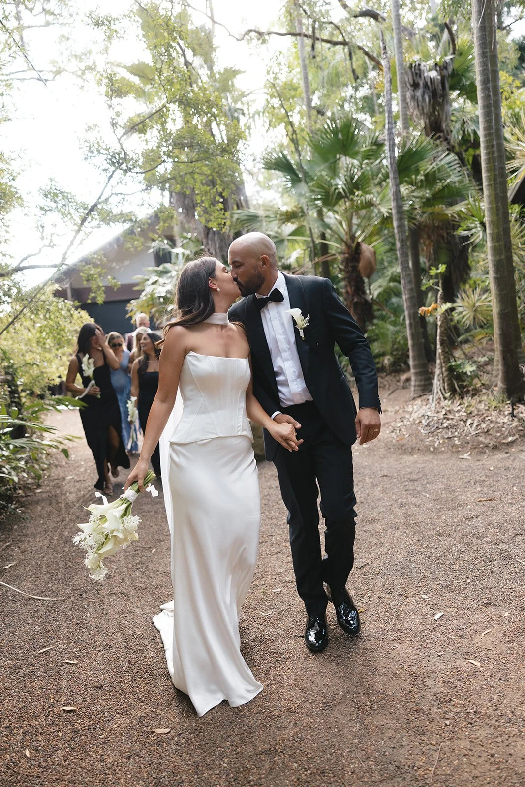 A newlywed couple, the bride in a white strapless wedding gown and the groom in a black tuxedo, kiss while walking hand-in-hand on a dirt path surrounded by lush greenery, with bridesmaids following behind.