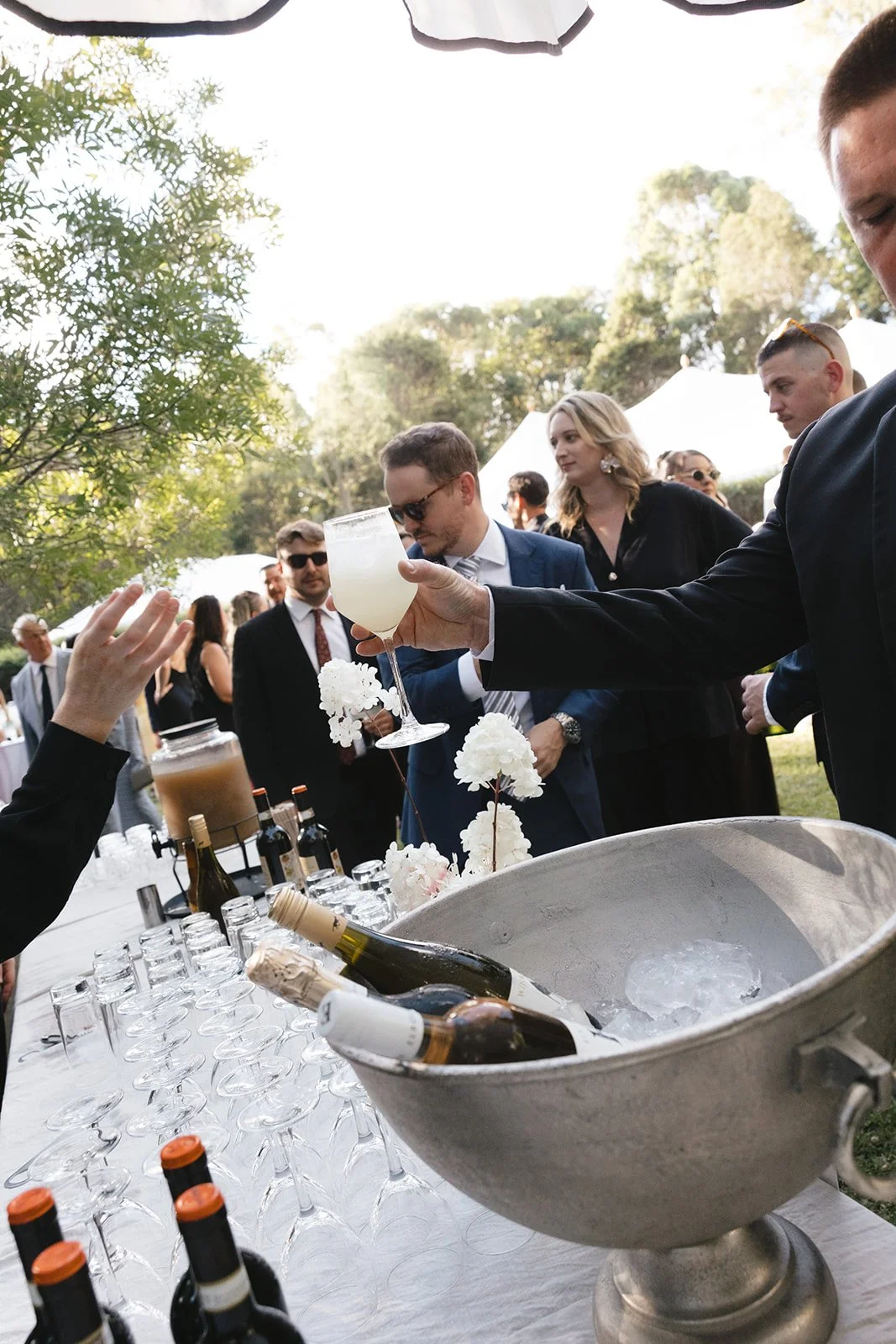 People at an outdoor event serving drinks from a large ice-filled bowl, with bottles of wine, glasses, and floral decorations on the table. A Hunter Valley Outdoor Wedding Event at Leaves and Fishes.