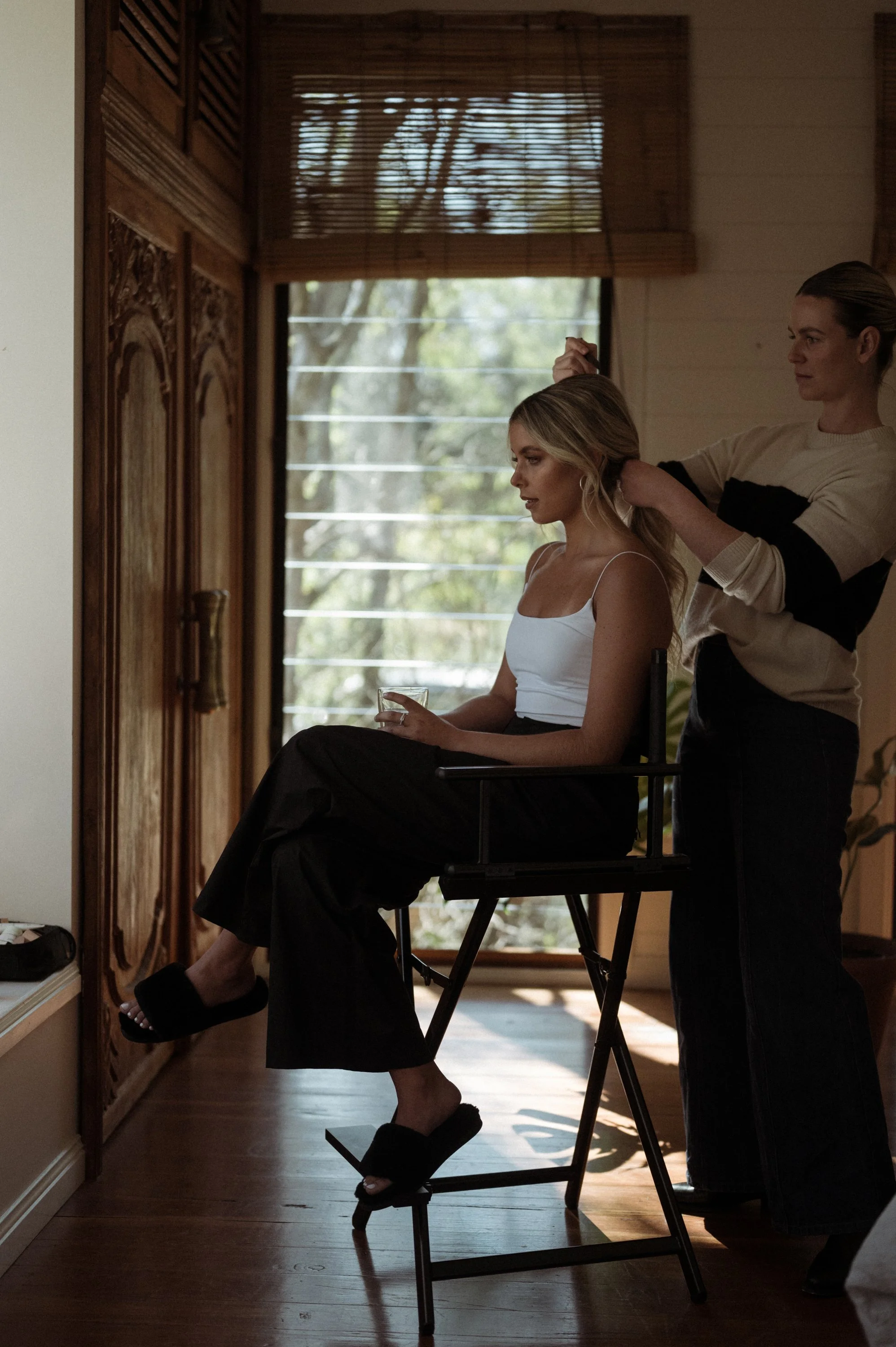 A woman is sitting on a chair while another woman styles her hair in a room with wooden walls and large windows with blinds.