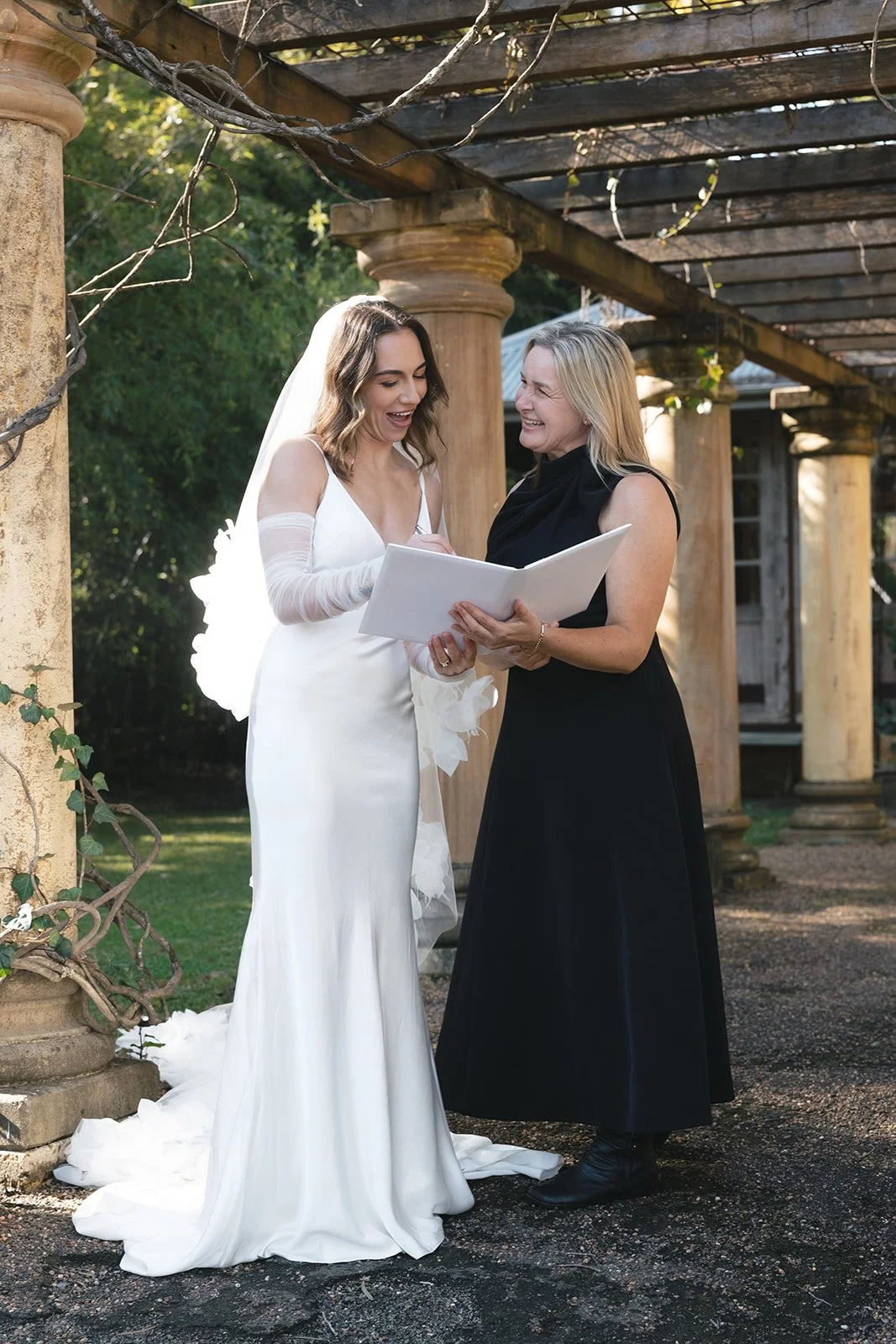 A bride in a white wedding dress and gloves shares a joyful moment with a woman in a black dress, holding a book outdoors under a pergola with stone pillars.
