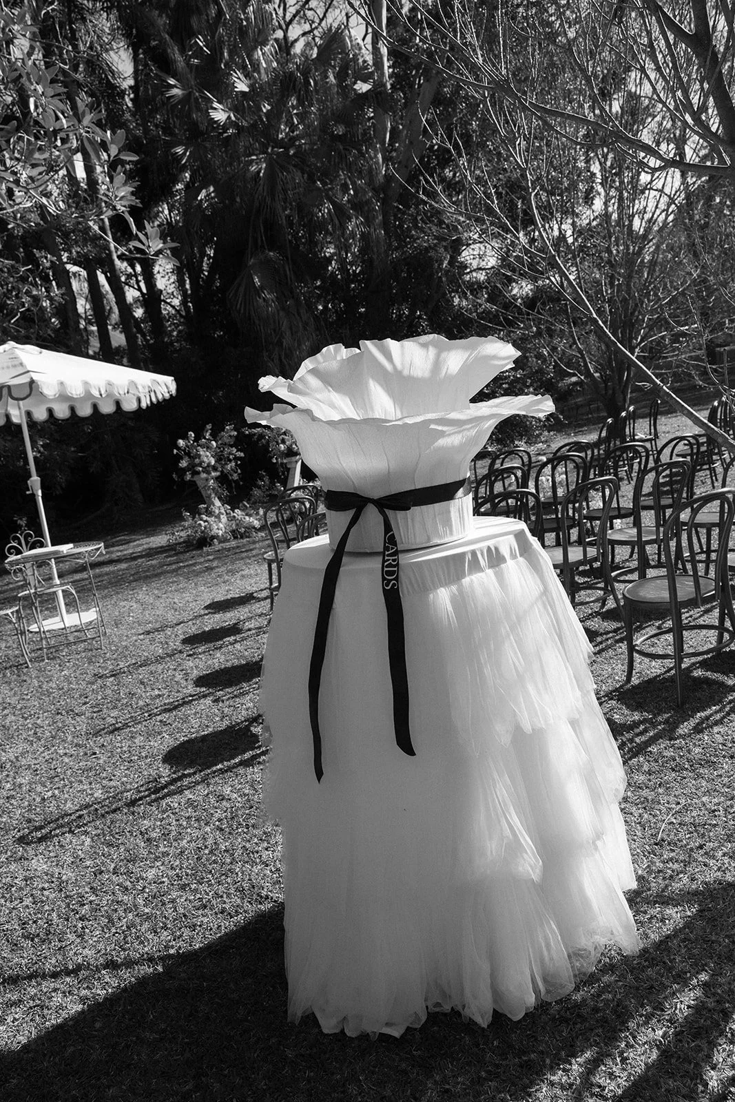 Decorative wedding arch with large white paper flowers, ribbons, and a tulle drape, outdoors on a grassy area with chairs arranged for an event and trees in the background.