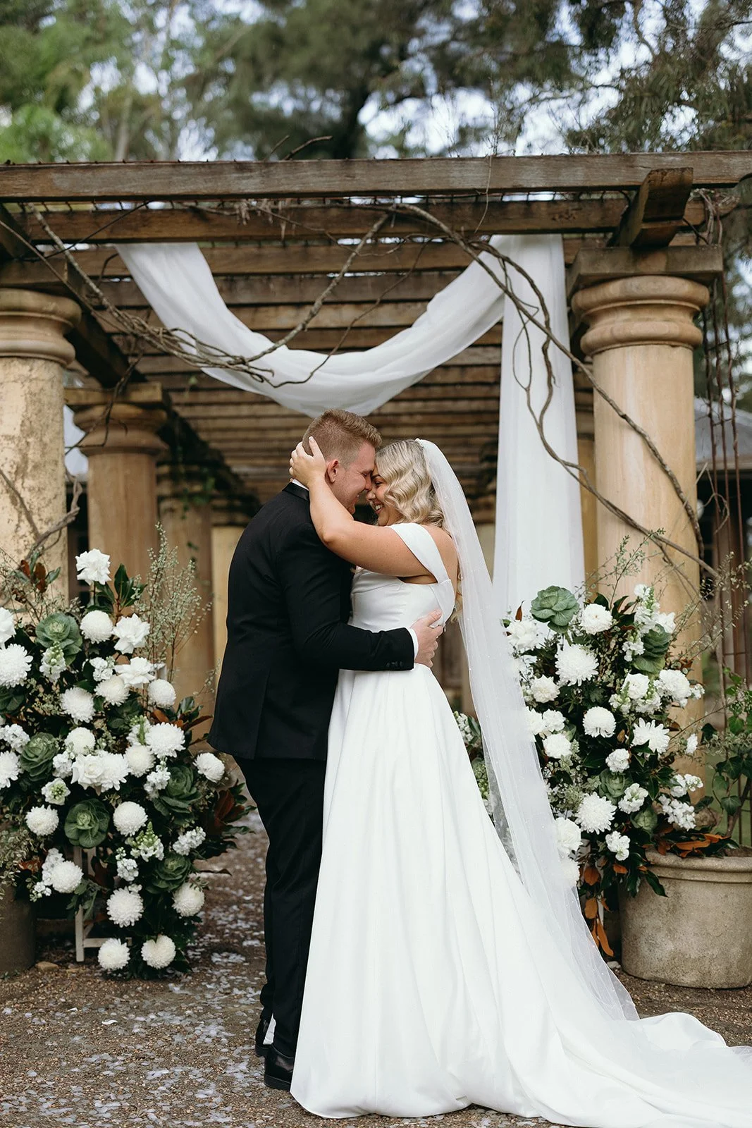 A newlywed couple sharing a kiss, standing close with foreheads touching, under a rustic wood and fabric wedding arch decorated with white flowers and greenery.