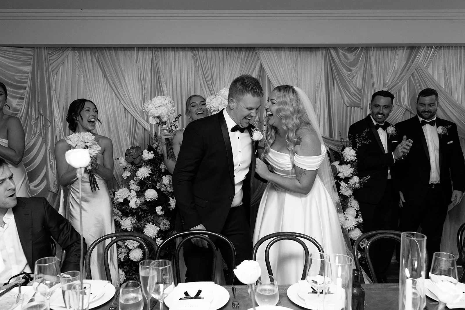 A black-and-white photo of a wedding celebration. The bride and groom are laughing and sharing a joyful moment at the center. The bride is wearing a wedding dress, and the groom is in a suit. Bridesmaids and groomsmen stand behind them, smiling and holding bouquets. The scene is decorated with flowers and draped fabric in the background.