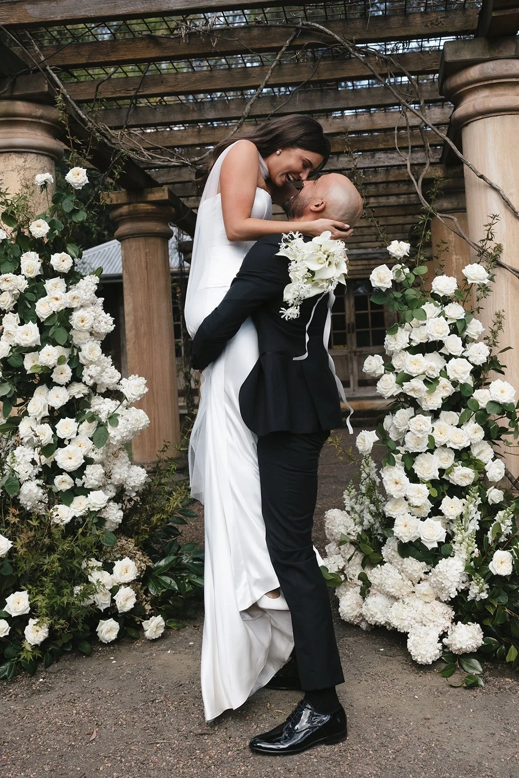 A bride and groom share a joyful moment under a wooden pergola decorated with white roses and greenery.