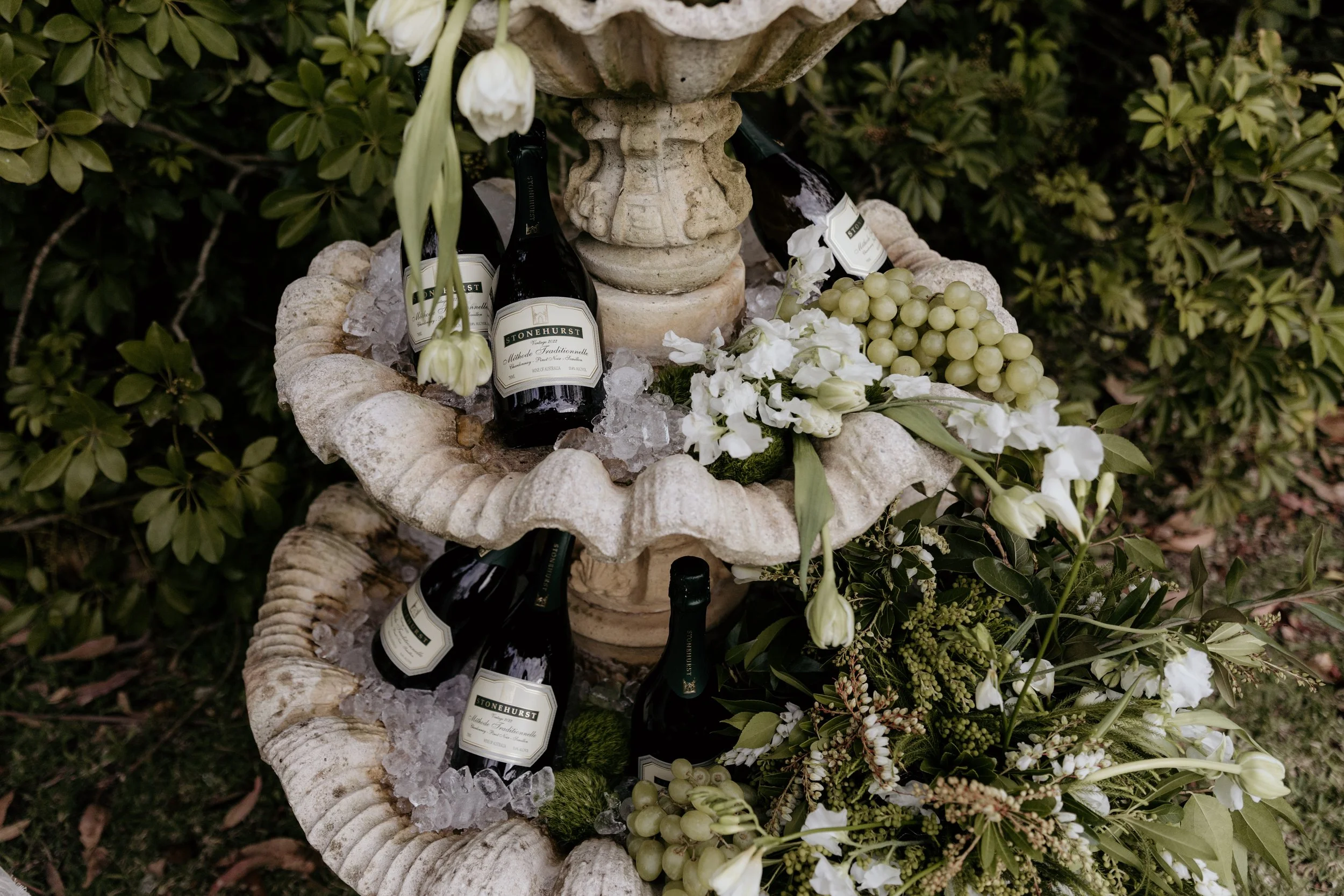 A two-tier stone fountain filled with ice, bottles of sparkling wine, and grapes, surrounded by white flowers and greenery.