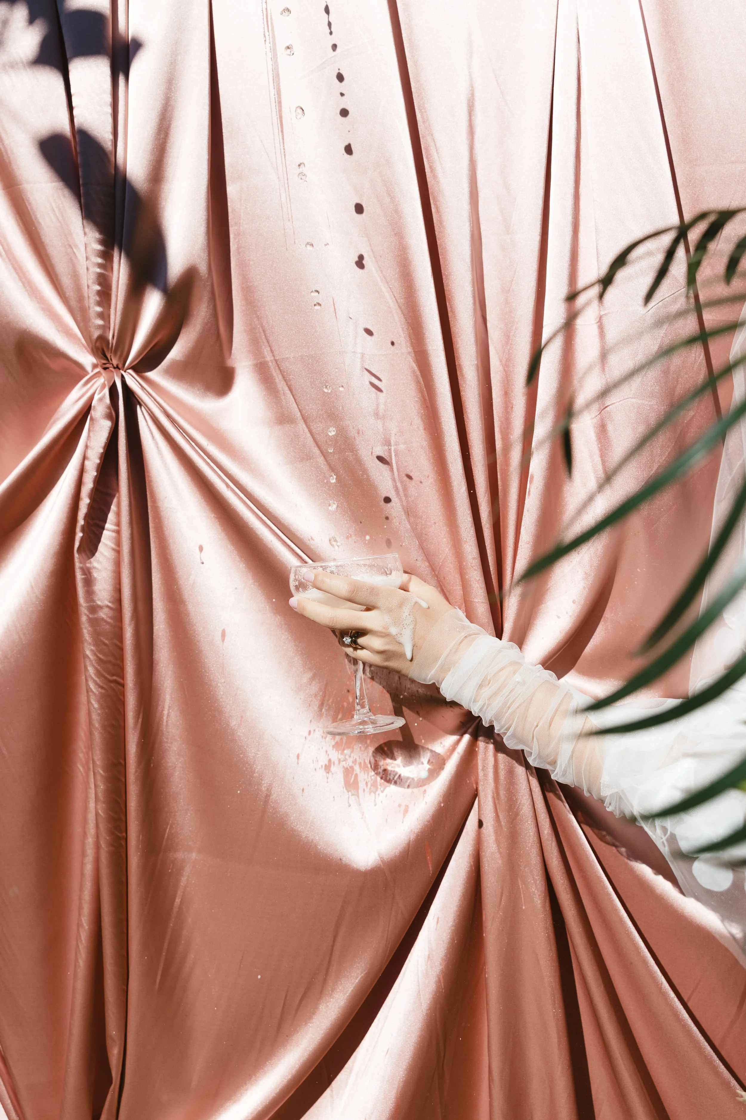 Person with a sheer glove spilling a drink on a pink satin backdrop.