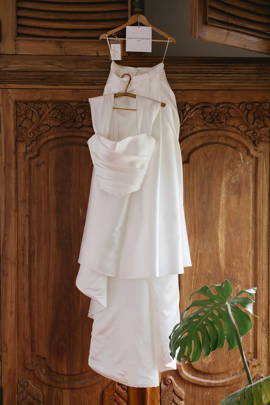 A white wedding gown and a matching veil hanging on wooden hangers against a carved wooden wardrobe. A note with 'Karen Willis Holmes' is attached to the hanger of the gown.