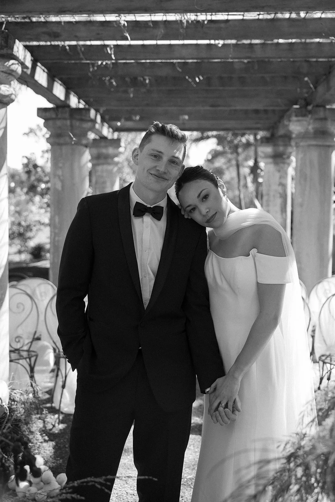 A black and white photo of a newlywed couple holding hands and smiling, with the woman resting her head on the man's shoulder, under a wooden pergola with stone columns, outdoor setting.