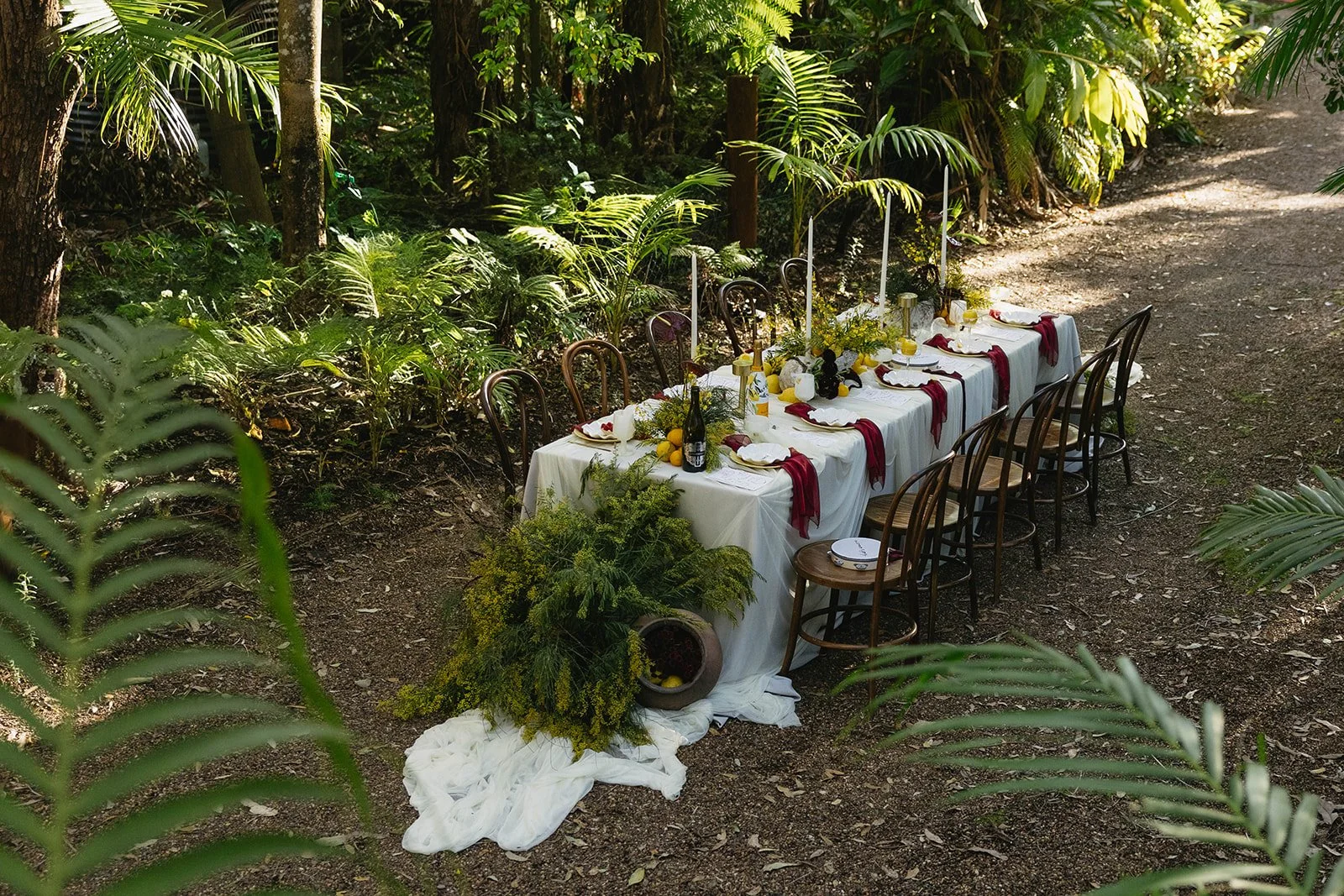 A long outdoor dining table set along a forest path, covered with a white tablecloth and decorated with candles, greenery, citrus fruit, and deep red napkins, surrounded by wooden chairs at Leaves and Fishes