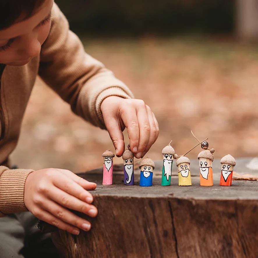 A child placing small painted wooden mushroom figures on a tree stump, each with a smiling face and colorful painted bodies at Secret Garden