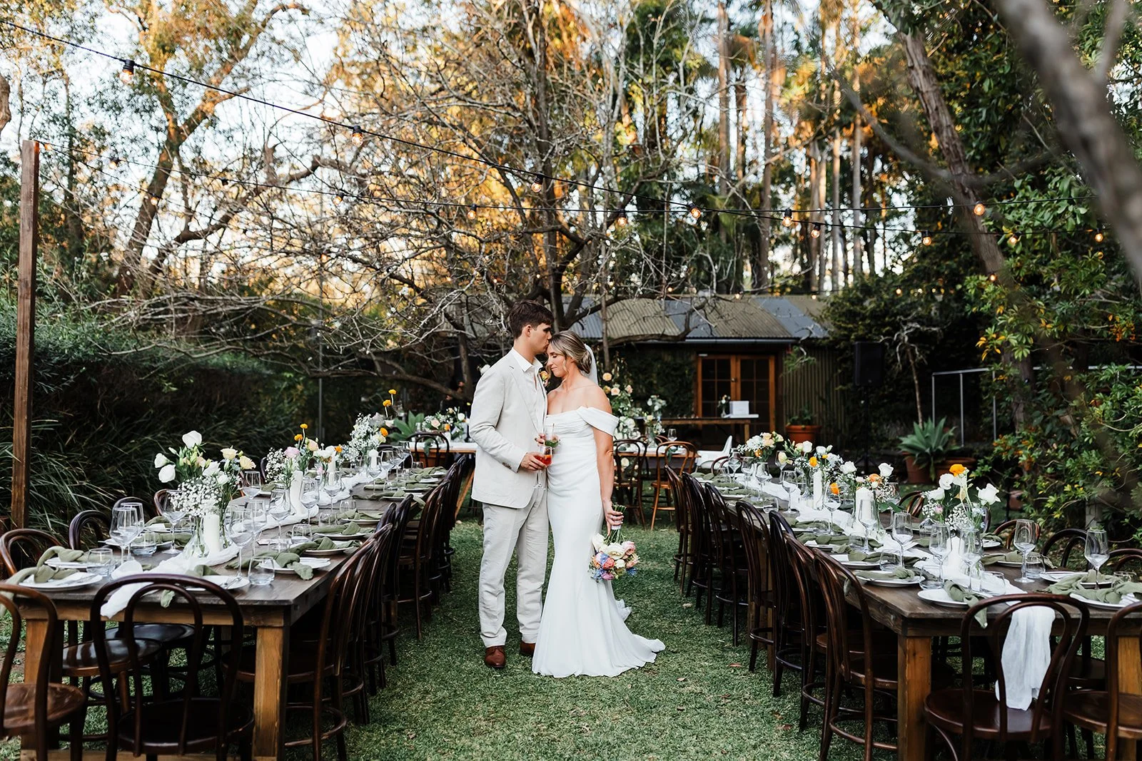 Couple standing at an outdoor garden wedding reception with long tables, flowers, and string lights at Leaves and Fishes