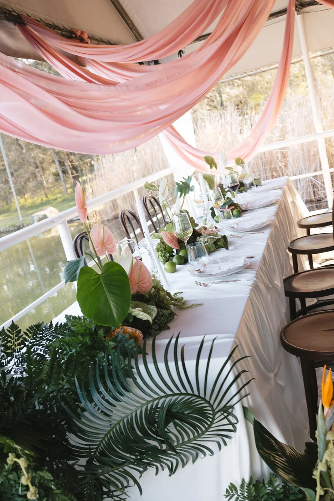 A decorated outdoor dining table with white tablecloth, floral centerpiece, glassware, and place settings under pink fabric drapes, overlooking a pond and greenery.
