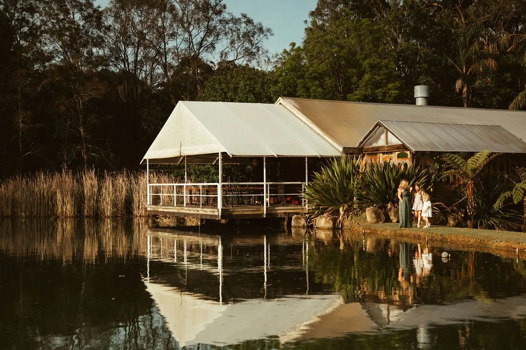 A lakeside scene featuring a house with a metal roof, a gazebo with a white canopy, and two women with a young girl near the water, surrounded by trees and plants at Leaves and Fishes
