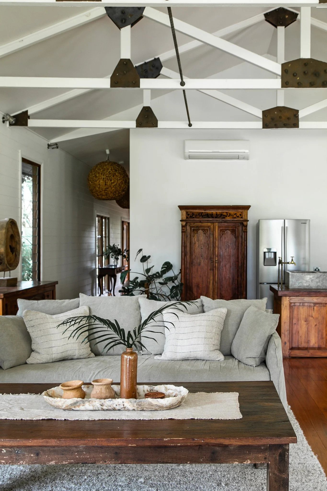 A cozy living room with a light-colored sofa, decorative pillows, a wooden coffee table with pottery and a vase with green leaves, wooden furniture, large windows, a stainless steel refrigerator, and a white wall-mounted air conditioner. The room has a high vaulted ceiling with exposed white beams and dark metal brackets.