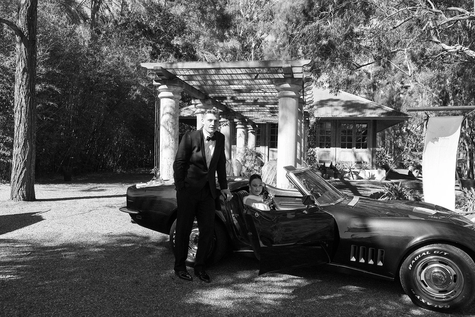 A black and white photo of a man in a tuxedo standing next to a woman in a dress seated in a vintage sports car, outdoors with trees and a building in the background.