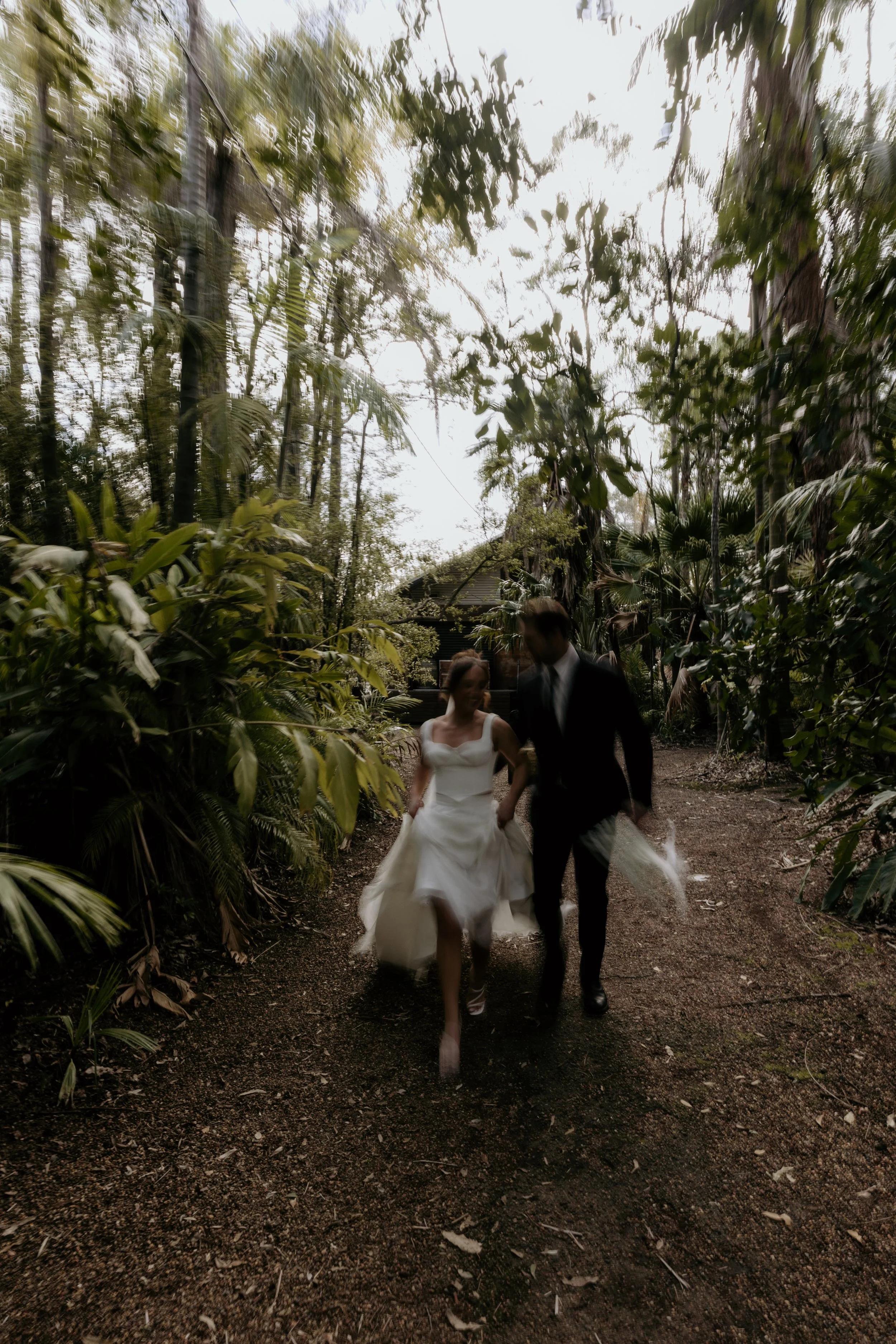 A bride and groom walking together on a forest trail during daytime, surrounded by lush green trees and foliage at Leaves and Fishes