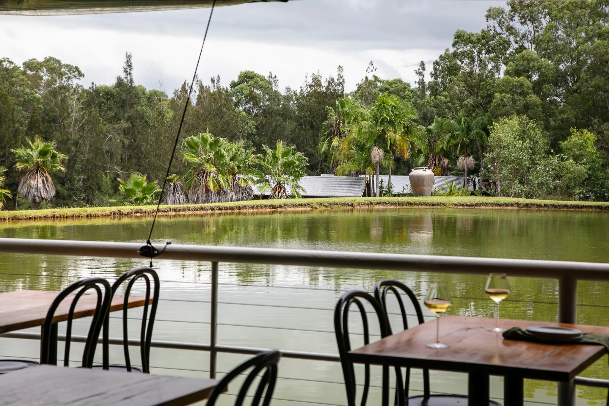 Dining area on a patio overlooking a pond with trees and tropical plants in the background, with two glasses of white wine on a table at The Pavillion