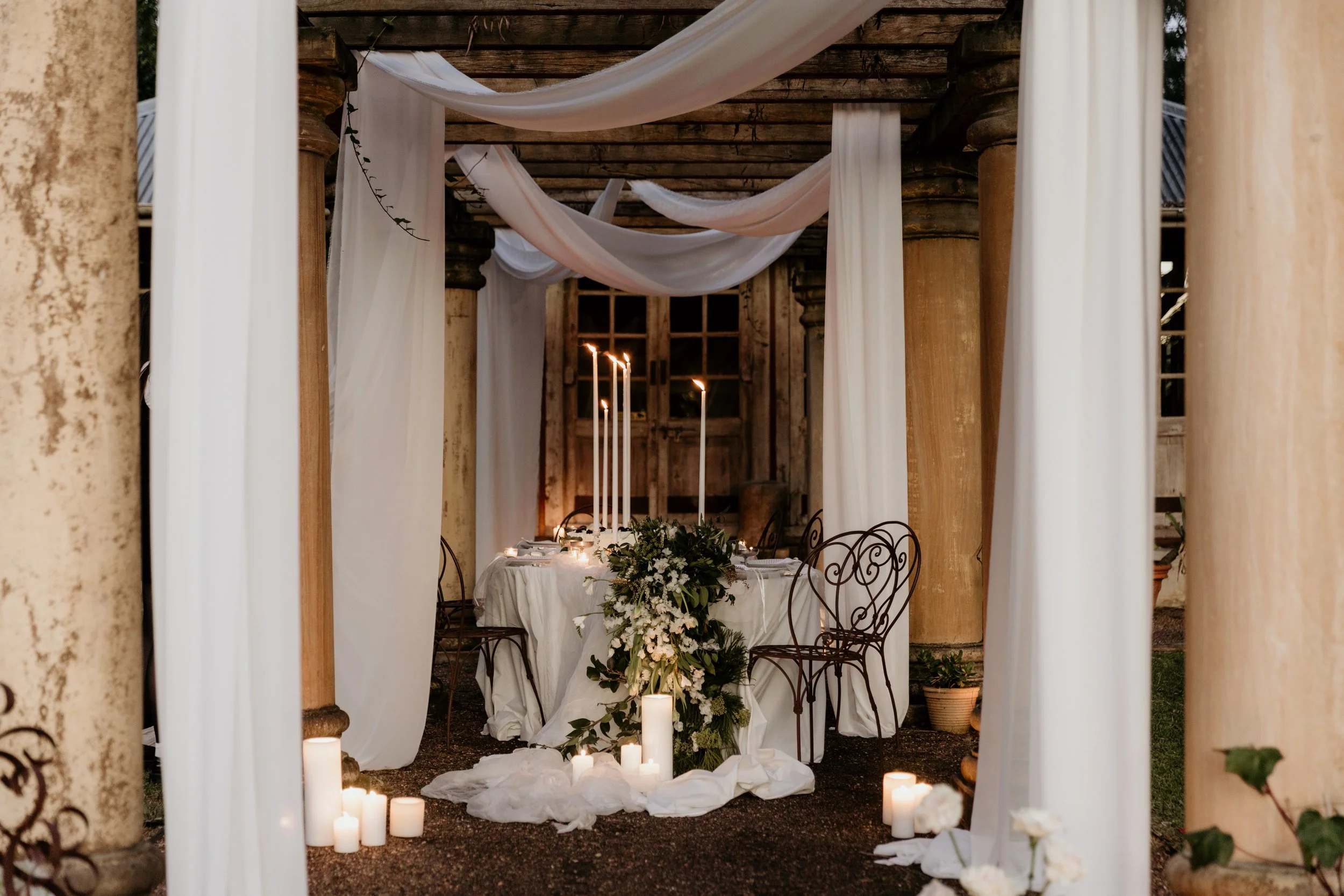 An elegant outdoor wedding altar with white drapery, candles, and floral arrangements, set against a rustic wooden structure with columns.