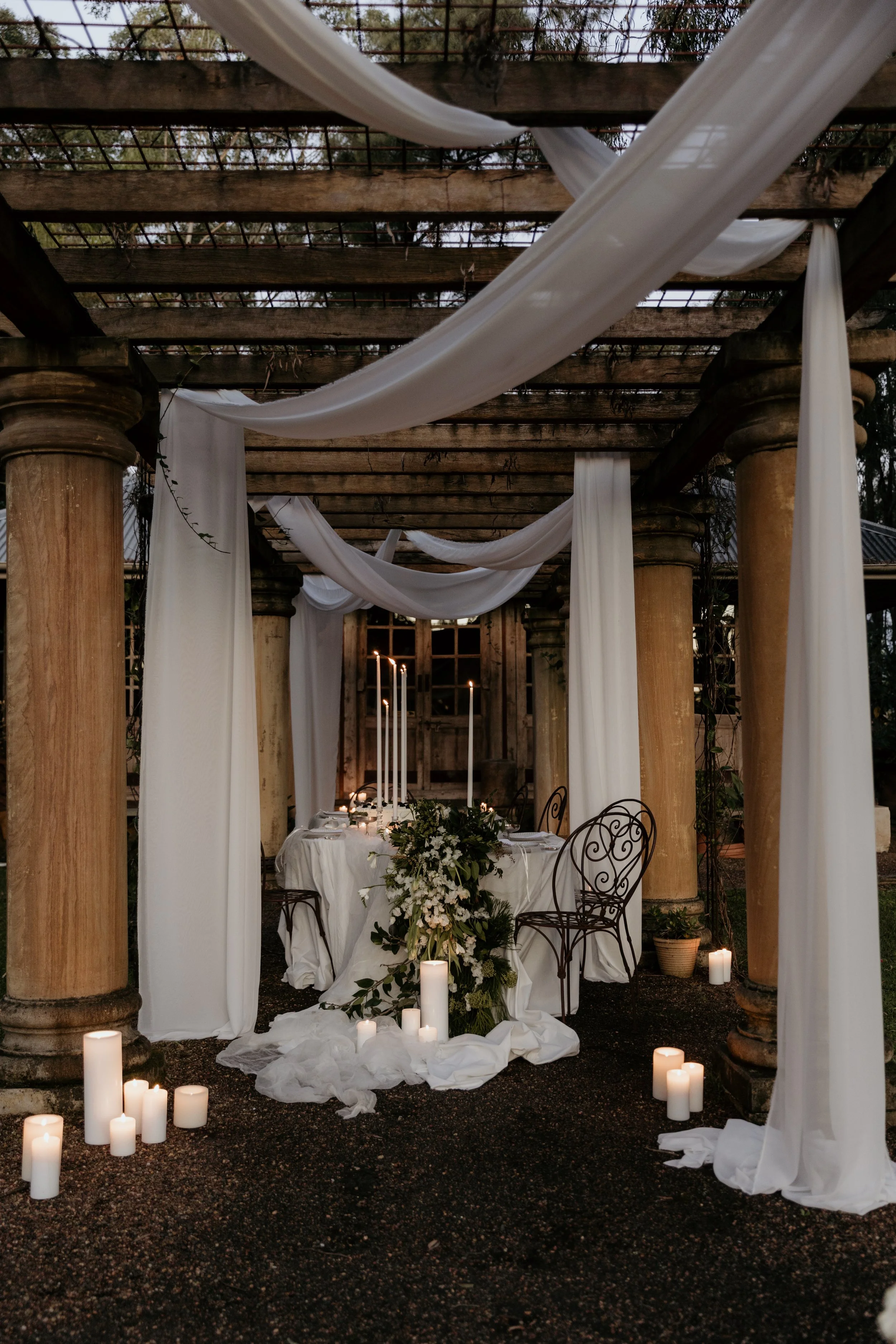 Elegant wedding decoration under a pergola with white drapery, candles, and a floral centerpiece.