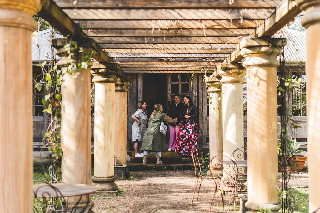 People greeting each other on a porch of a rustic wooden house with columns, surrounded by outdoor furniture and greenery at Leaves and Fishes.
