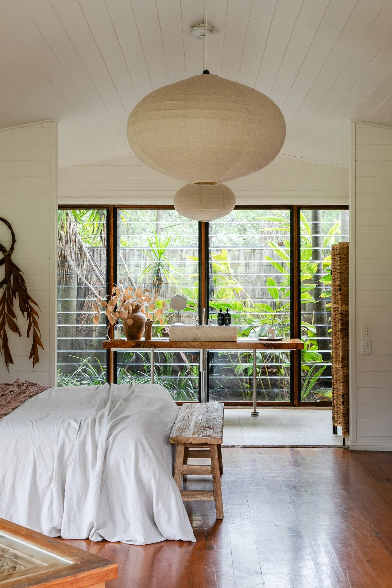 An interior view of a room with a bed covered in white linen, a wooden bench, and a large paper lantern hanging from the ceiling. There are large windows with greenery visible outside, and a vanity table with vases and bottles.