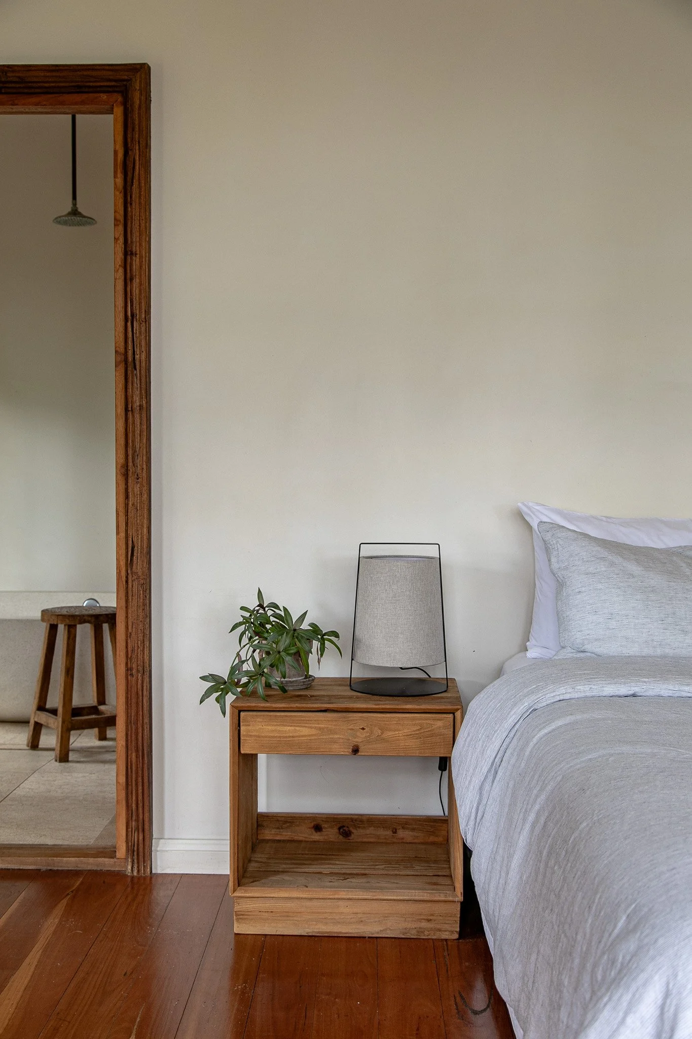 Bedroom scene with a wooden nightstand holding a potted plant and a modern lamp, next to a bed with light gray bedding. A doorway with a wooden frame leads to a bathroom with a showerhead and a stool at Leaves and Fishes