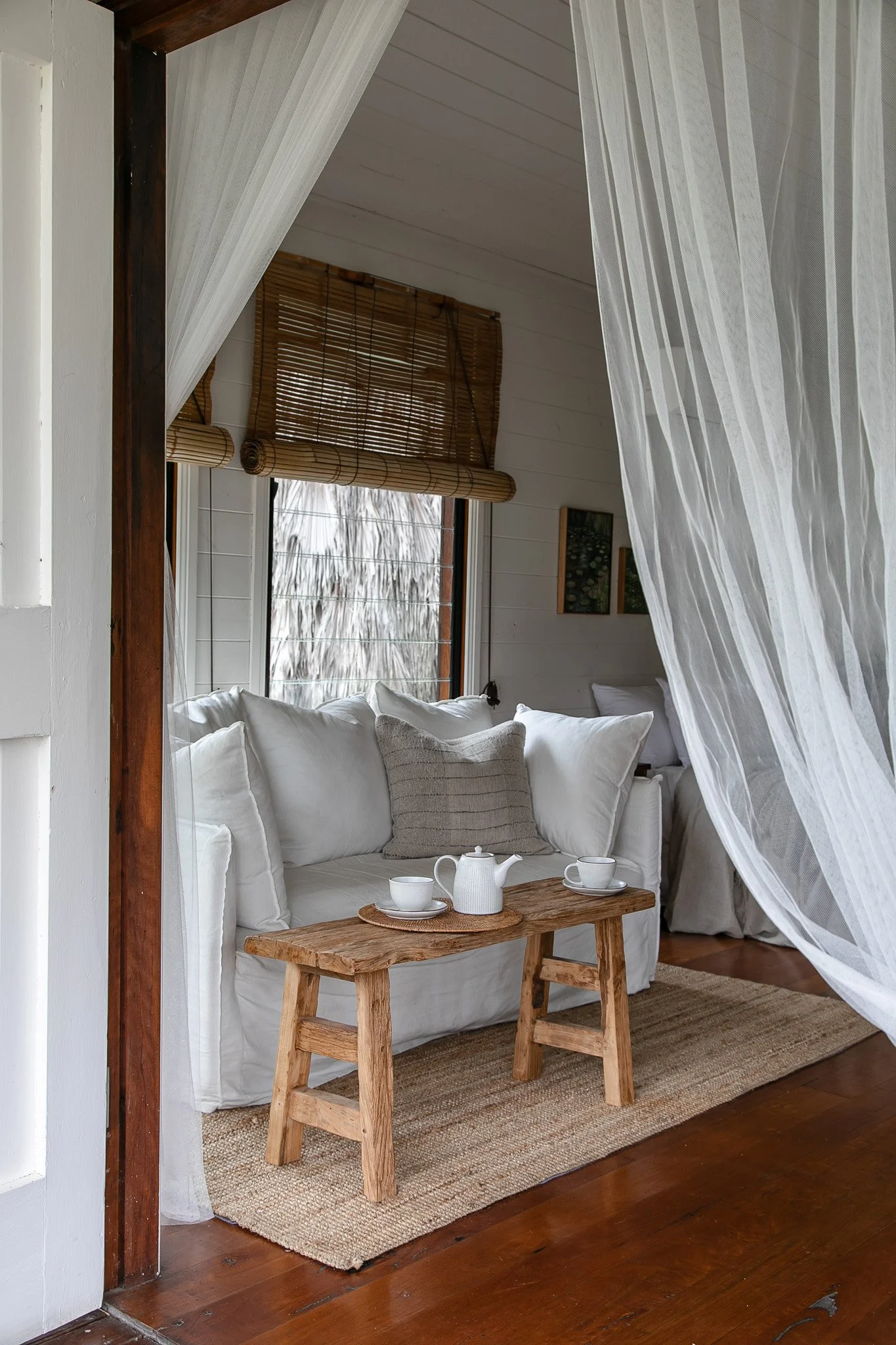 Cozy living room with white sofa, wooden coffee table set for tea, window with bamboo blinds, and sheer curtains.