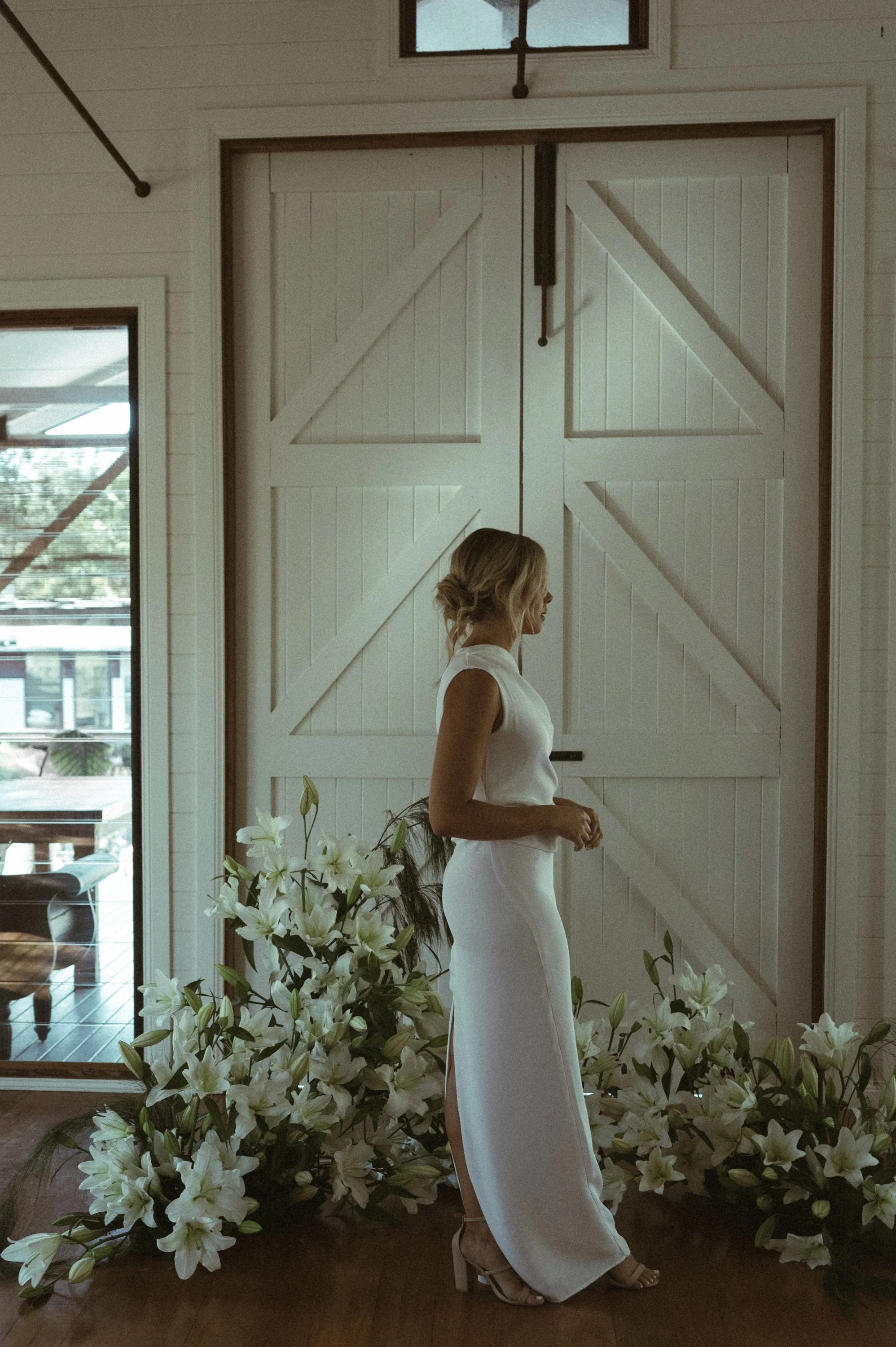 A woman in a sleeveless white dress and heels standing indoors beside white lilies, with large barn-style doors behind her and windows letting in natural light.