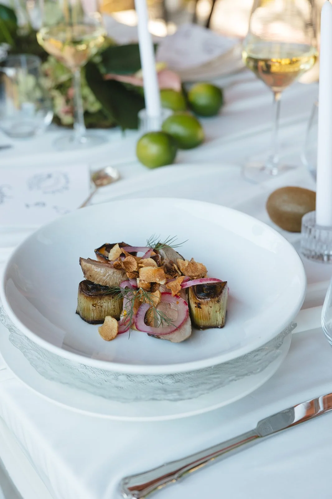 A plated dish of grilled or roasted vegetables, garnished with herbs and crispy toppings, set on a white table with wine glasses, green limes, and candles in the background.