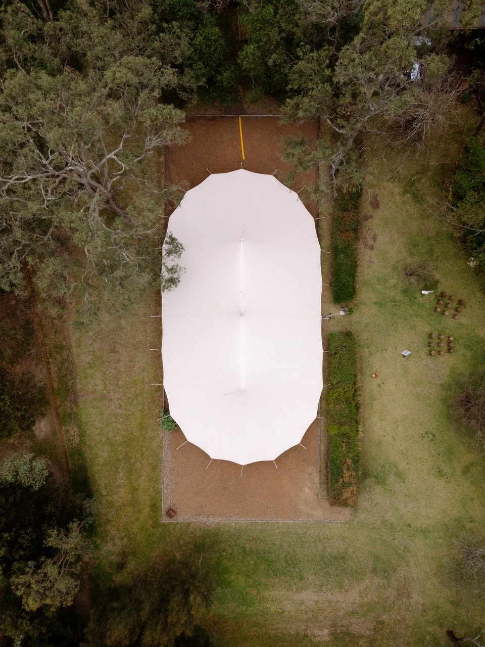 An aerial view of an outdoor white tent set up on a dirt area, surrounded by grass and trees.