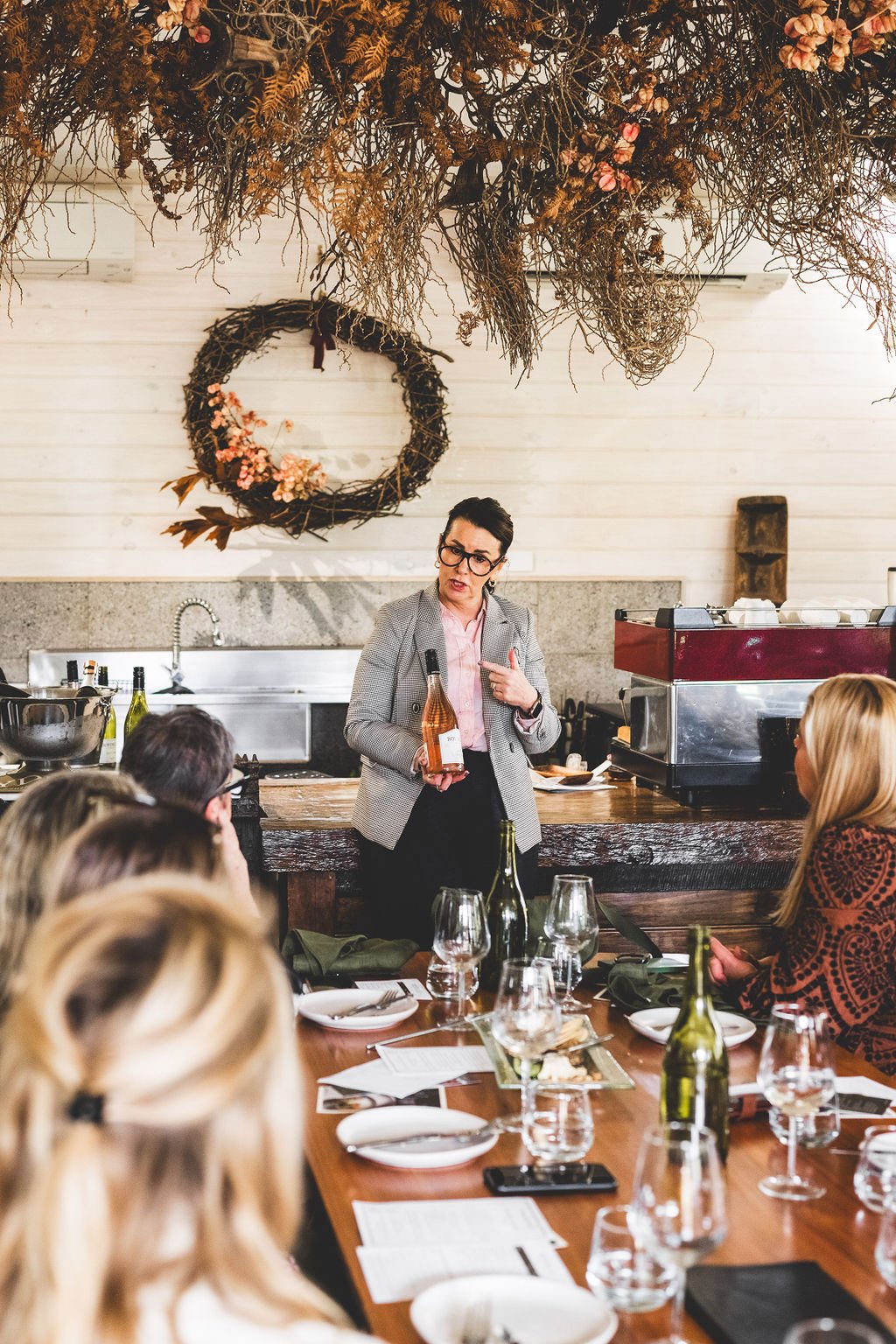 A woman giving a wine presentation to a group of people in a restaurant or winery setting, with decorative dried floral arrangements and a wine wreath hanging on the wall behind her at Leaves and Fishes