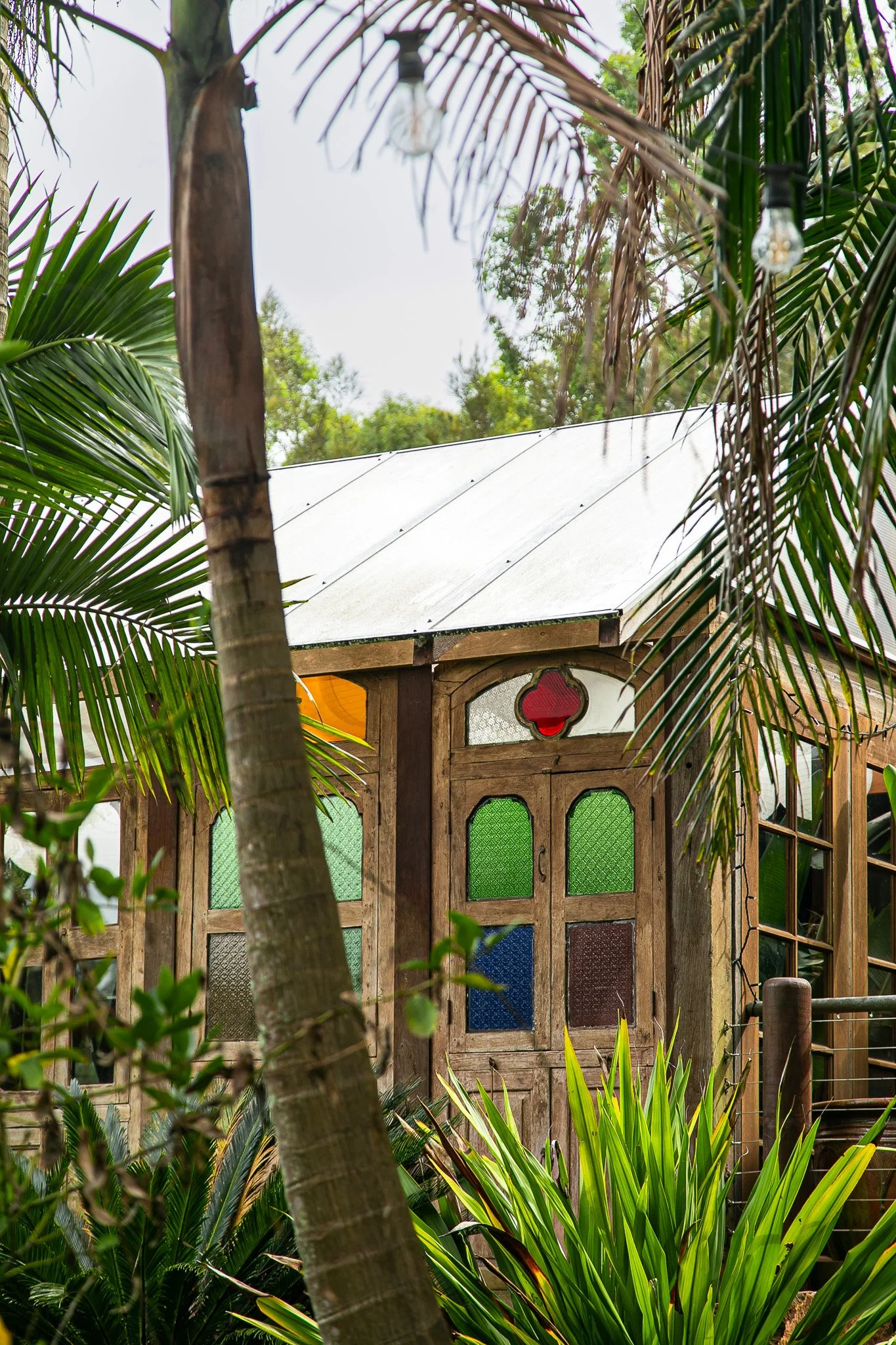 A small wooden structure with colorful stained glass windows surrounded by tropical plants and trees at Leaves and Fishes