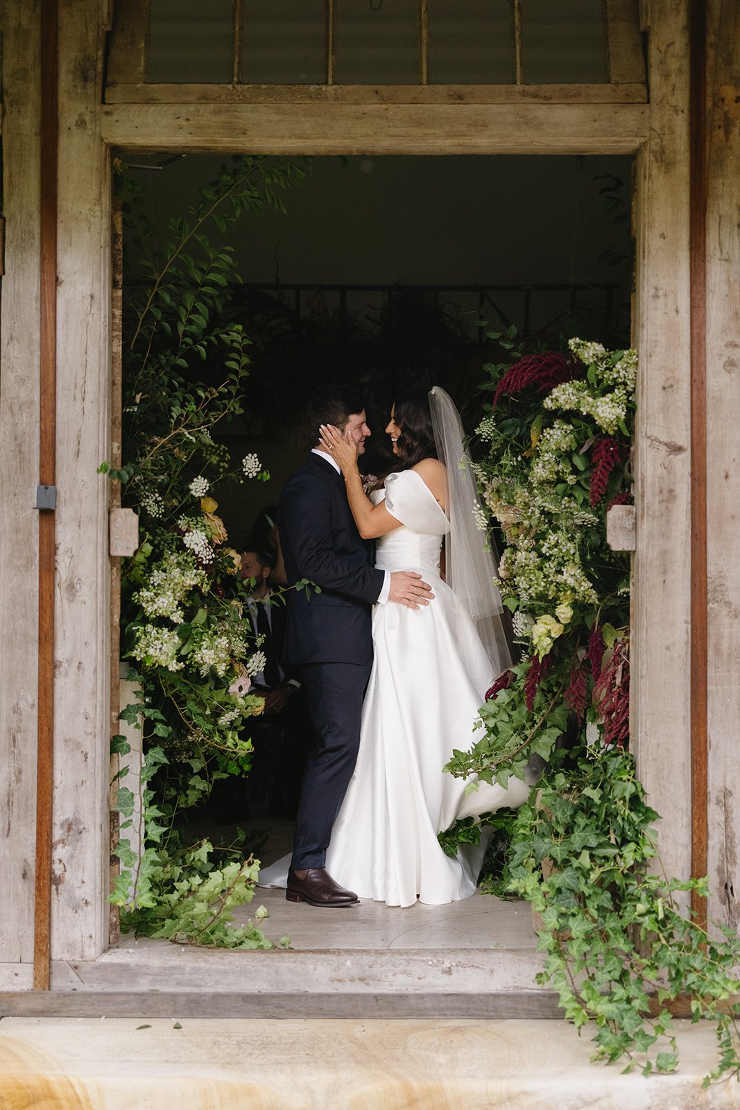 A bride and groom share a moment under a rustic wooden arch decorated with lush greenery and white flowers, during their wedding ceremony at Leaves and Fishes