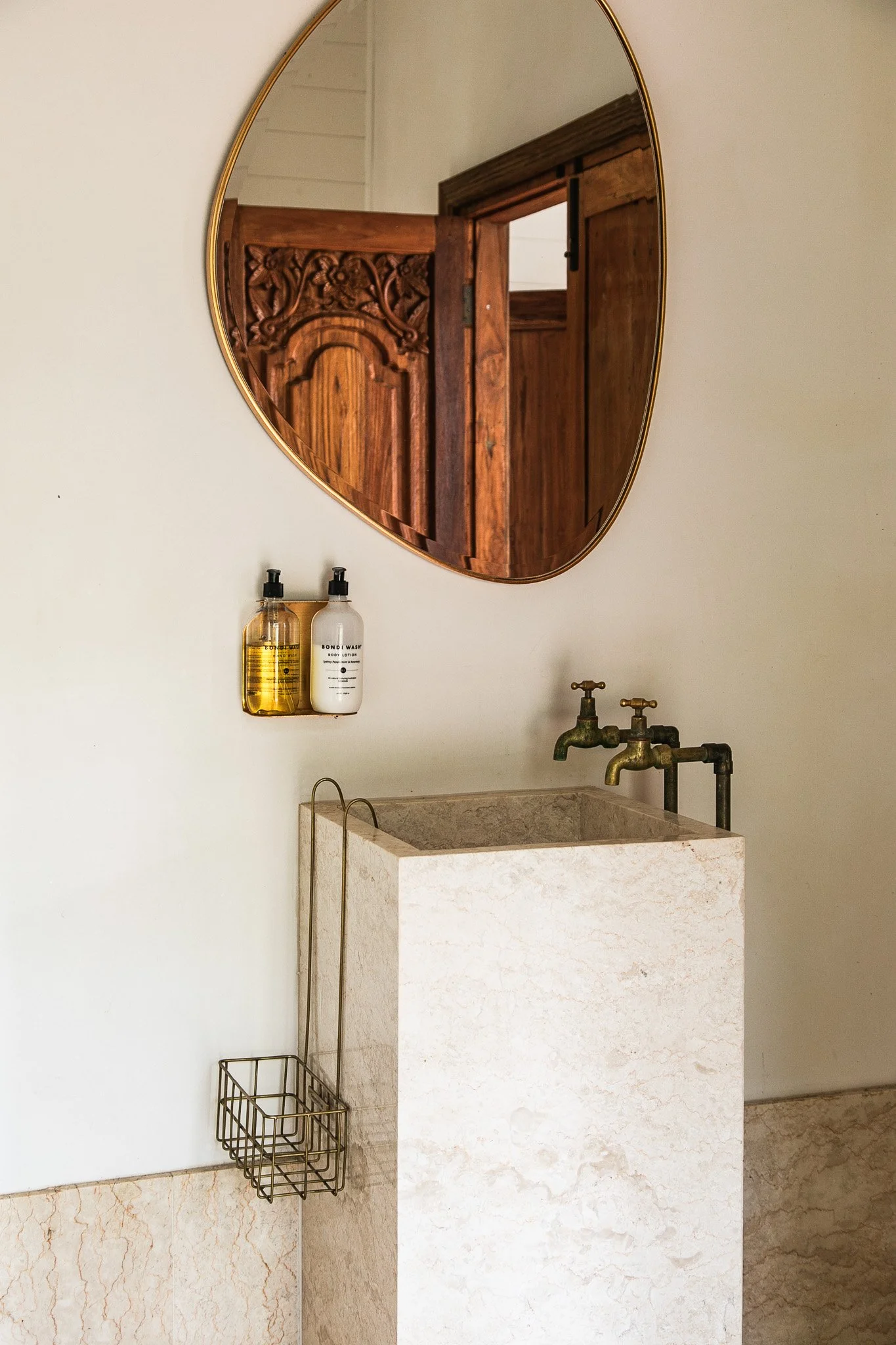 A bathroom corner with a white rectangular stone sink with vintage brass faucets, a round mirror with a gold frame, a wall-mounted soap dispenser holding two bottles, and a small wire basket hanging on the wall at Leaves and Fishes