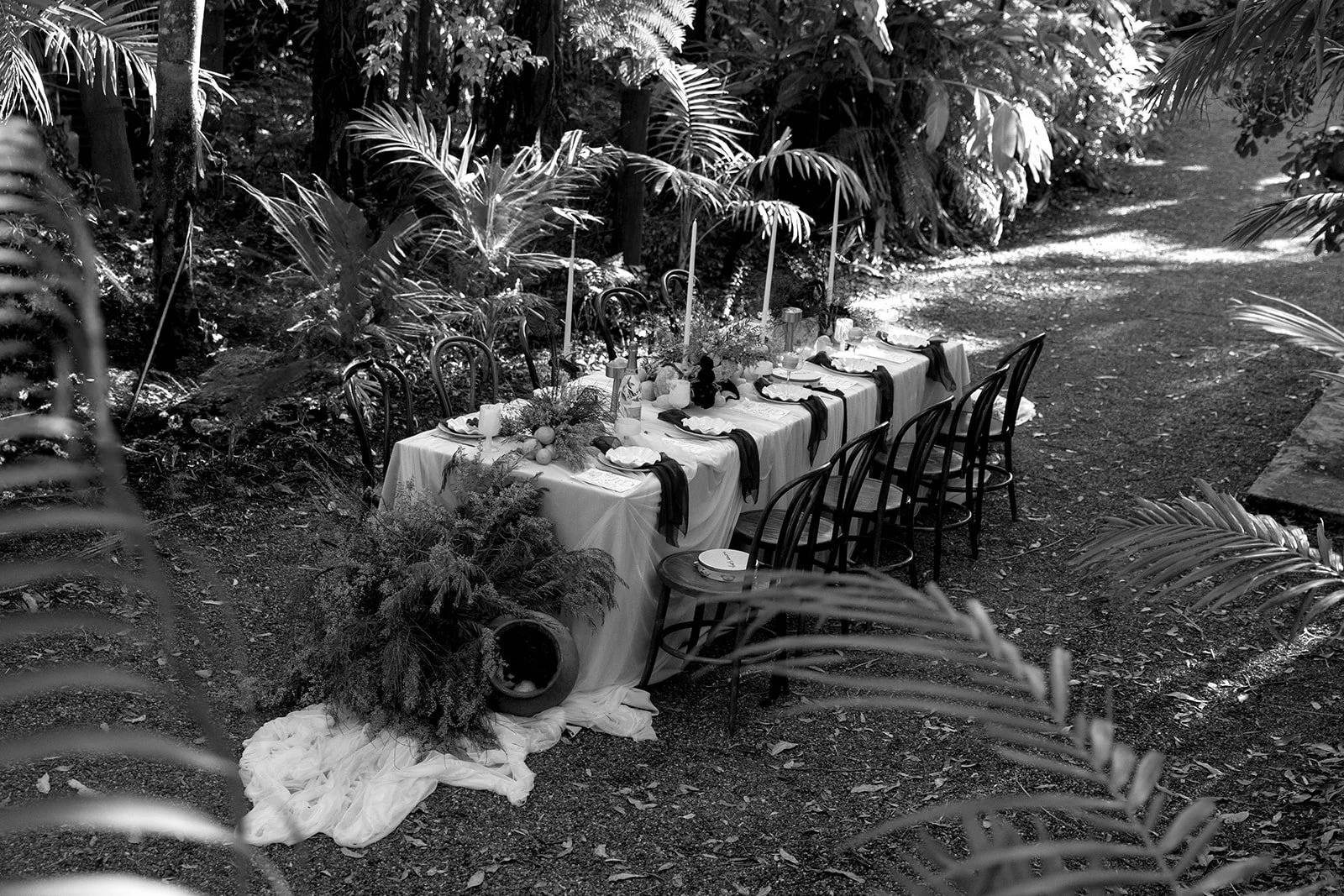 A long banquet table set with plates, glasses, and dip candles in a lush outdoor tropical garden with tropical plants and trees.