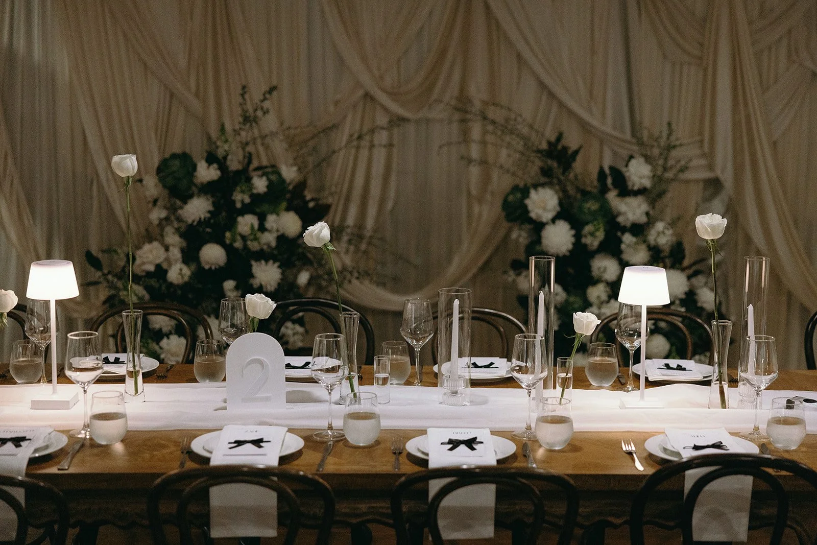 Elegant banquet table set with white flowers in tall stems, white candles, wine glasses, plates with black ribbon napkins, and small lamps, with floral arrangements and draped curtains in the background.