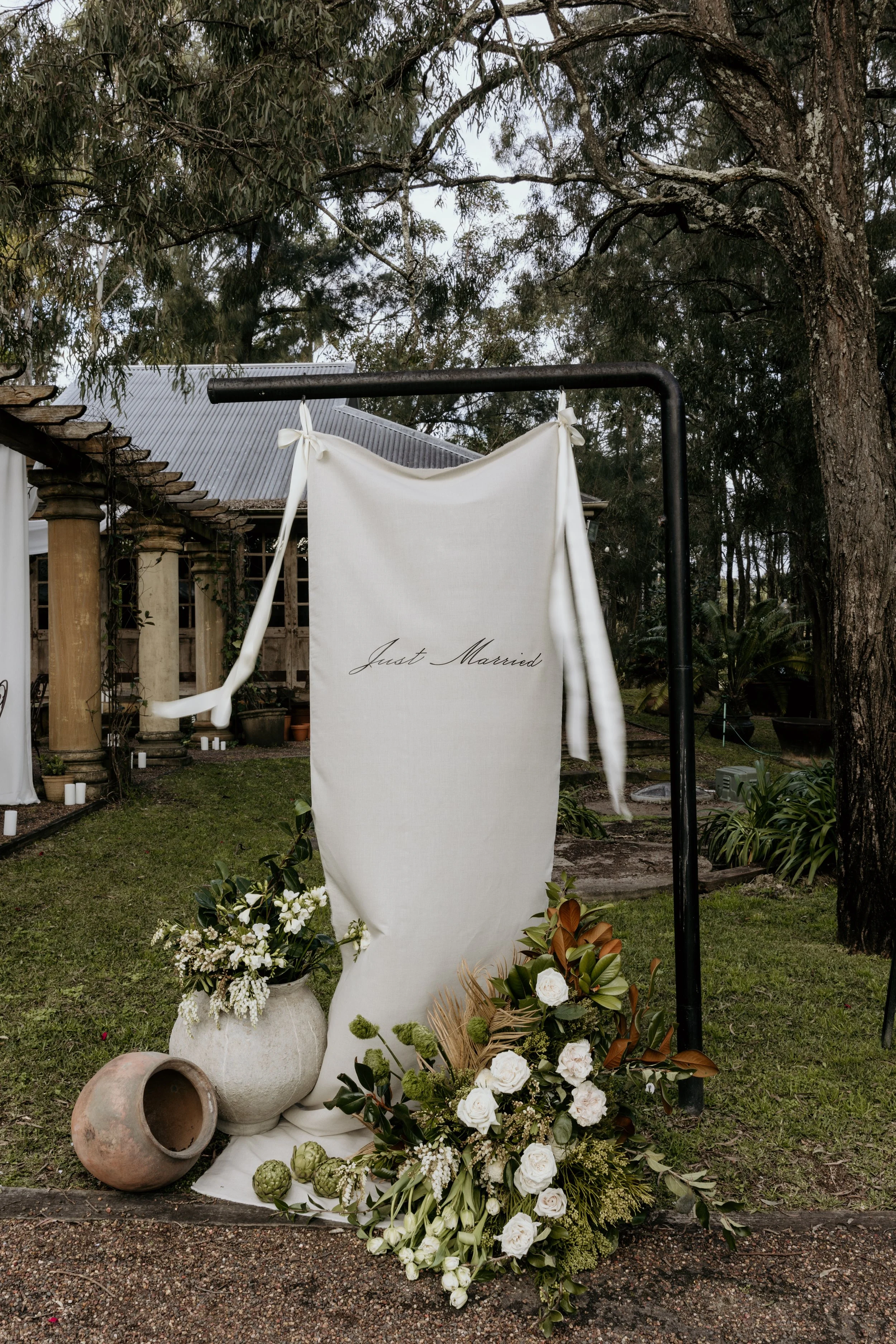 Outdoor wedding decor with a fabric banner reading 'Just Married,' surrounded by white flowers and pottery vases, set in a garden with trees and a building in the background.