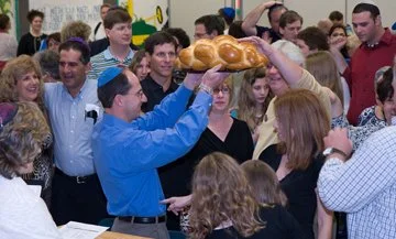 A man in a blue shirt is holding a large challah bread as it is passed over a crowd during a celebration.