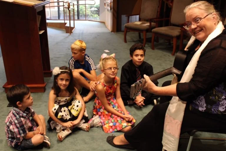 An elderly woman sitting on a chair playing an electric guitar, surrounded by five children sitting on the carpeted floor, in an indoor setting with chairs and a door in the background.