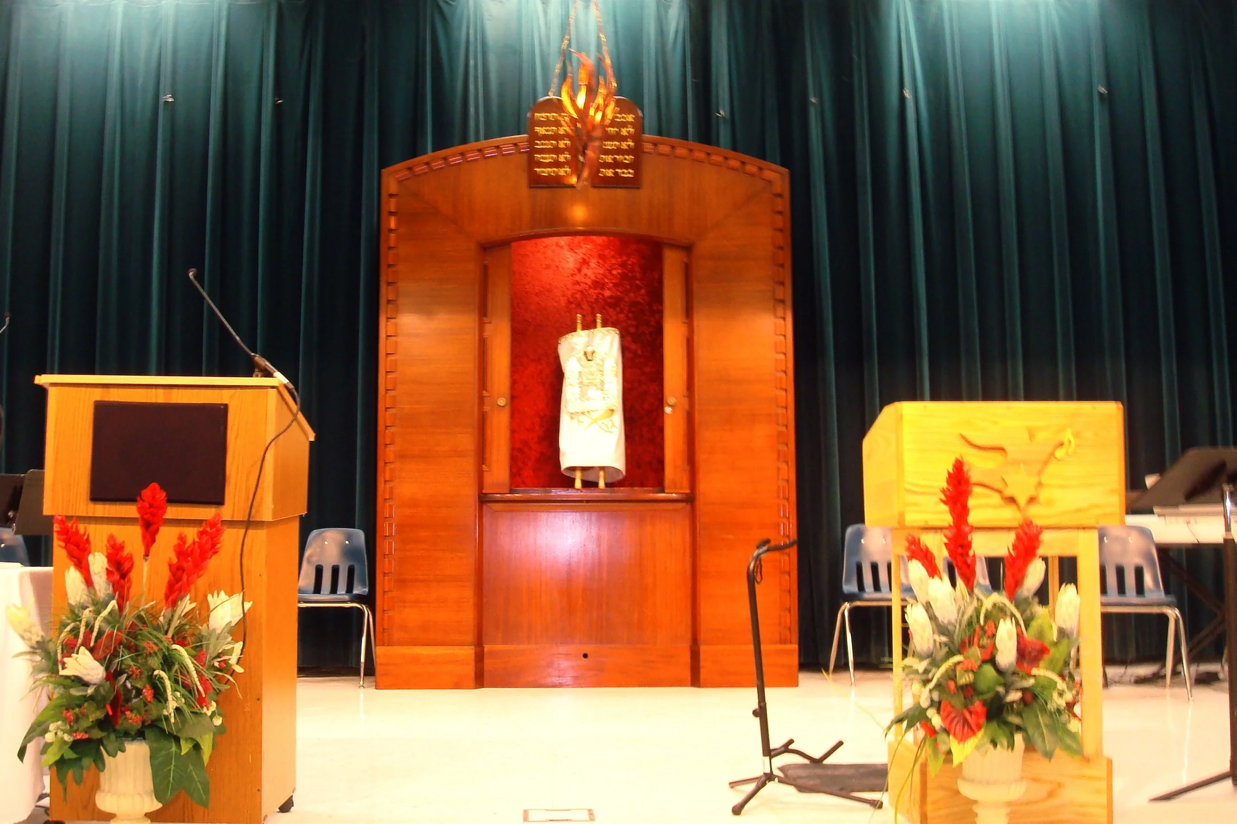 Interior of a synagogue with a Torah ark in the background, flanked by two wooden lecterns, decorated with colorful flower arrangements, and green curtains behind the ark.