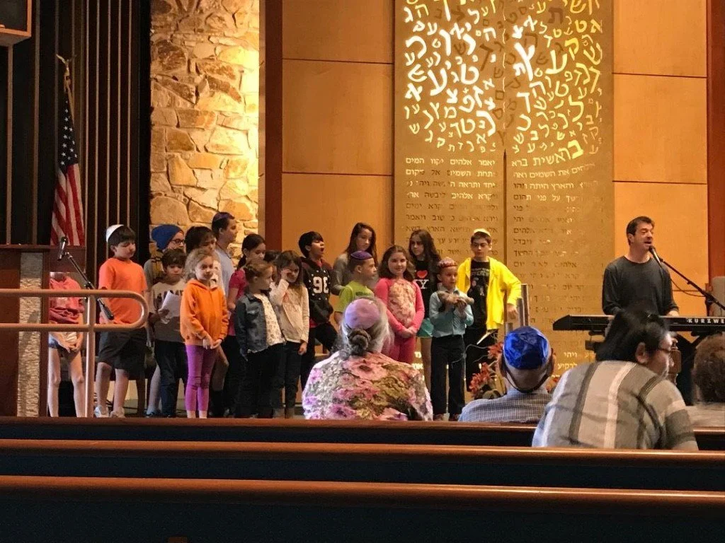 Children standing on stage in a church, singing or performing, with an adult man playing keyboard and singing. Audience members seated facing the stage, including a woman with a white headscarf and a man wearing a blue yarmulke. The background features a large wall with Hebrew inscriptions and a decorative illuminated tree symbol.