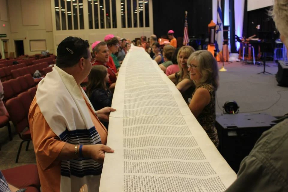 People participating in a Jewish tradition, holding a large Torah scroll during a religious or cultural event in a hall with stage and musical instruments.
