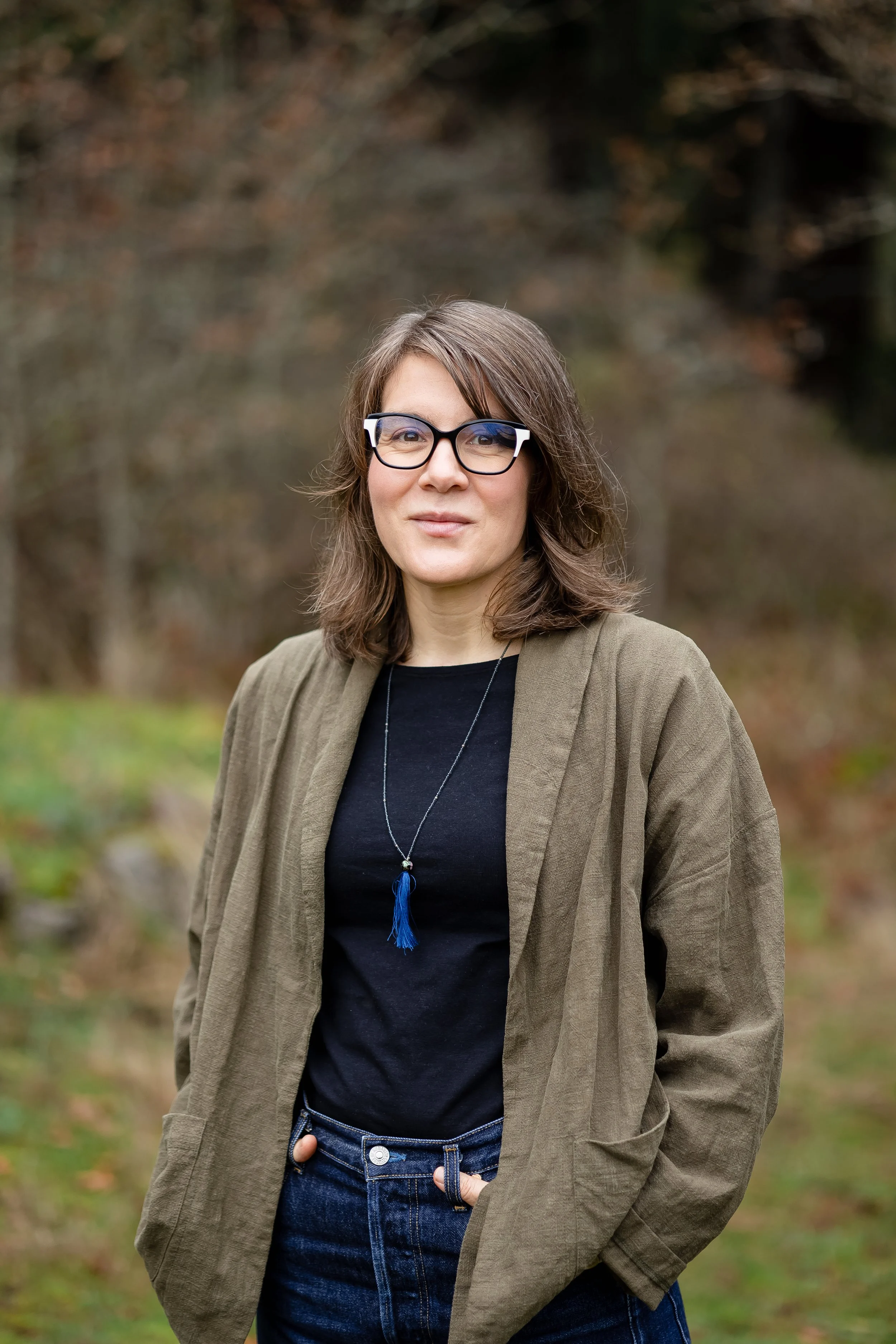 Smiling woman standing outside with hands in pockets, glasses, and brown hair wearing a casual green blazer and jeans; greenery fills the background.