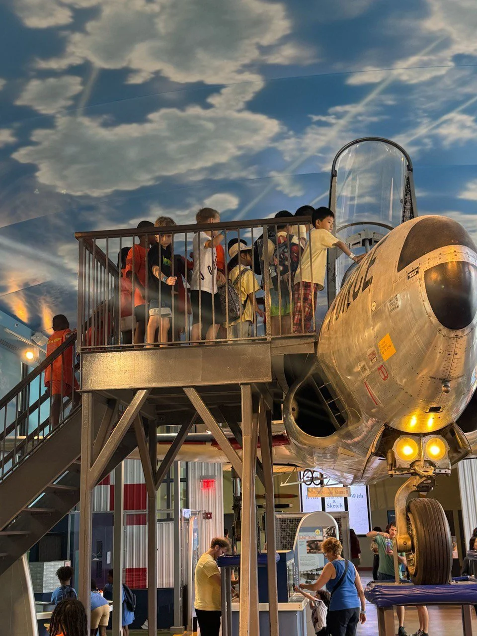 Children riding a vintage aircraft exhibit at a museum, with sky graphic display on the ceiling.