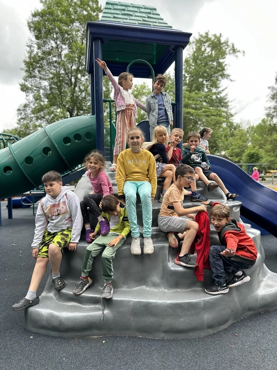 Children playing on a gray playground slide with a green tube tunnel, some sitting and others standing or climbing, under an overcast sky with trees in the background.