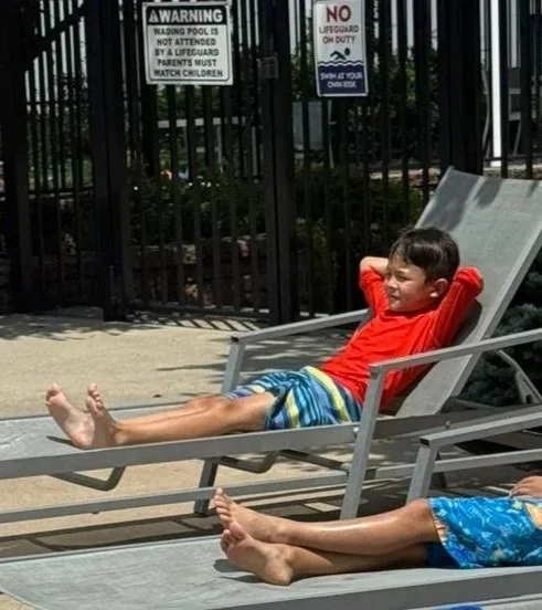 A young boy in a red shirt and colorful shorts relaxing on a lounge chair at a pool area.