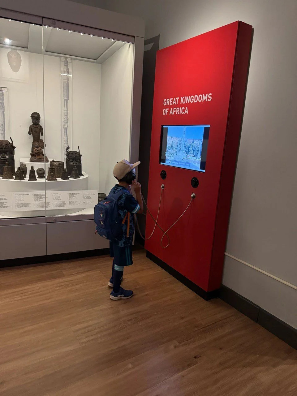 A young boy with a backpack and hat looking at a display in a museum titled 'Great Kingdoms of Africa', featuring artifacts and a screen showing African images.