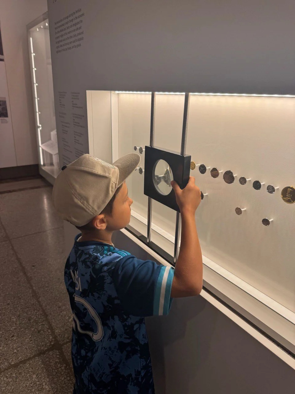 A young boy wearing a beige cap and a blue patterned sports jersey looking through a magnifying glass at a display of coins in a museum or exhibit.