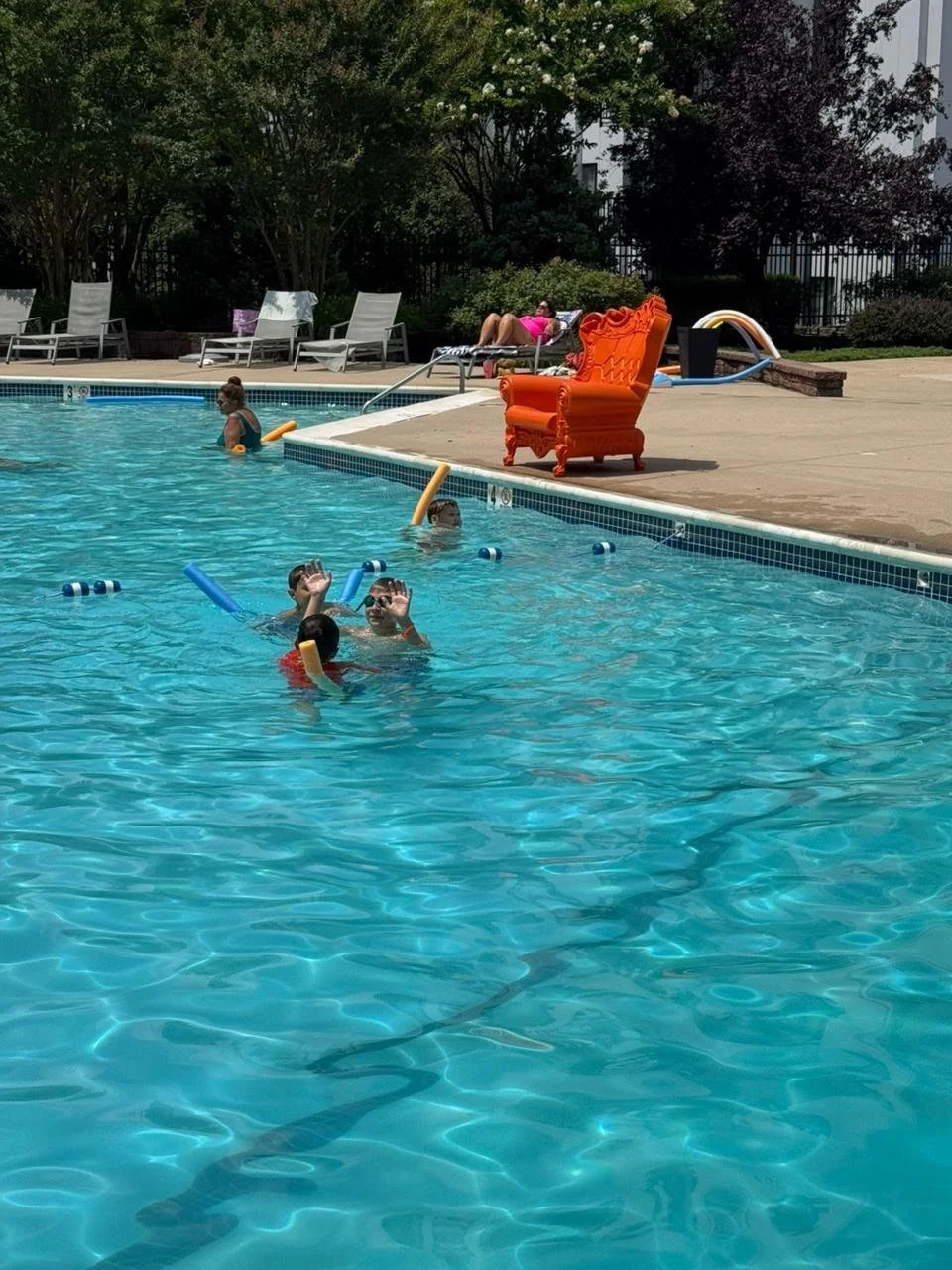 Kids swimming and playing with pool noodles in a swimming pool at a leisure area, with adults relaxing on lounge chairs and an orange ornate throne chair on the pool deck.